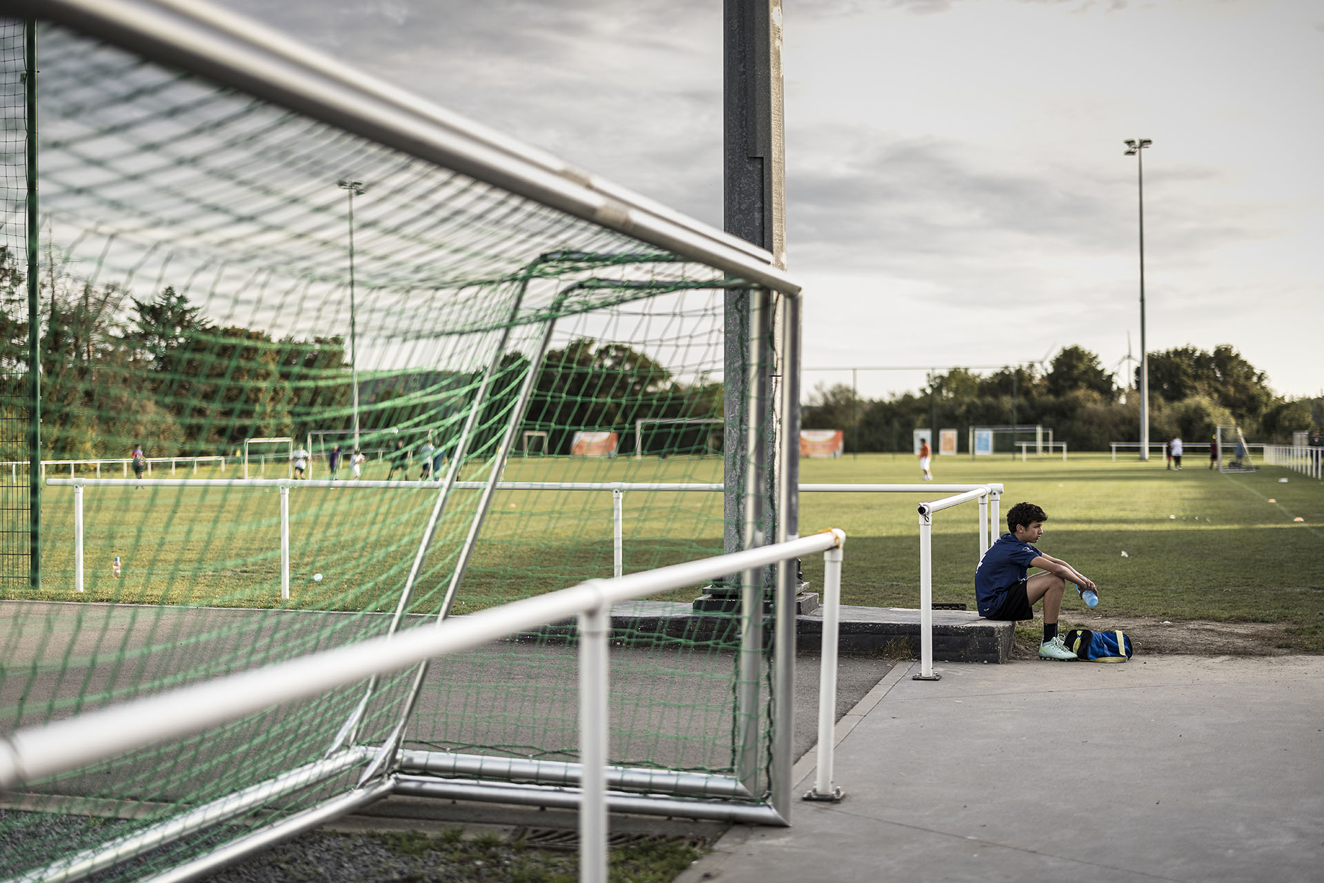 Fußballplatz mit einem Tornetz im Vordergrund; rechts sitzt ein Junge am Rand neben seiner Sporttasche.